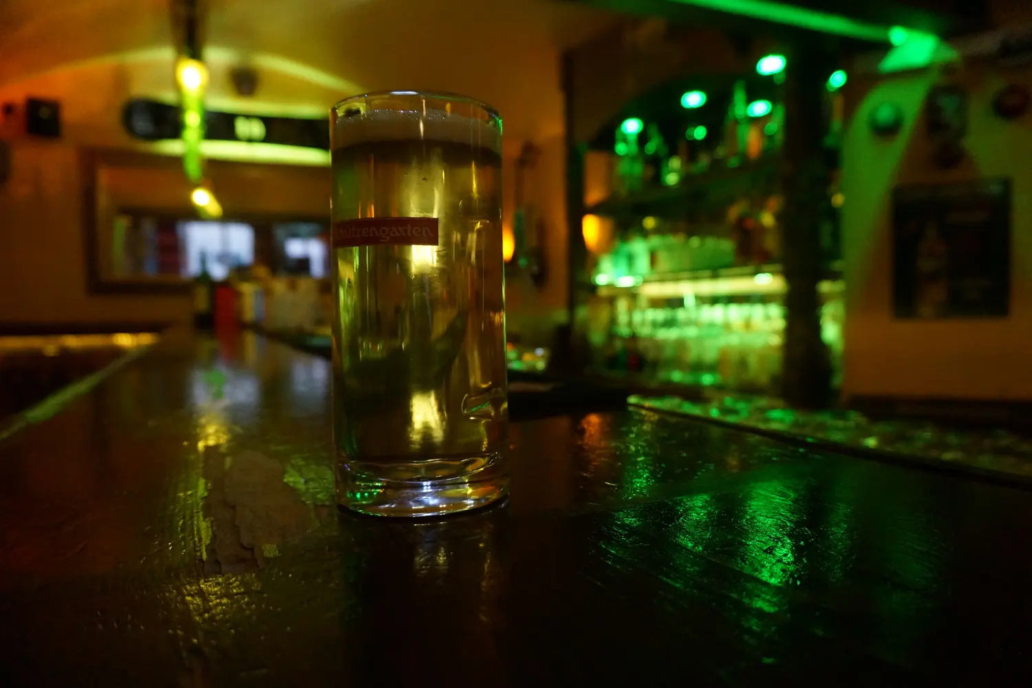 A full glass of beer on the counter, green light and the long bar behind it