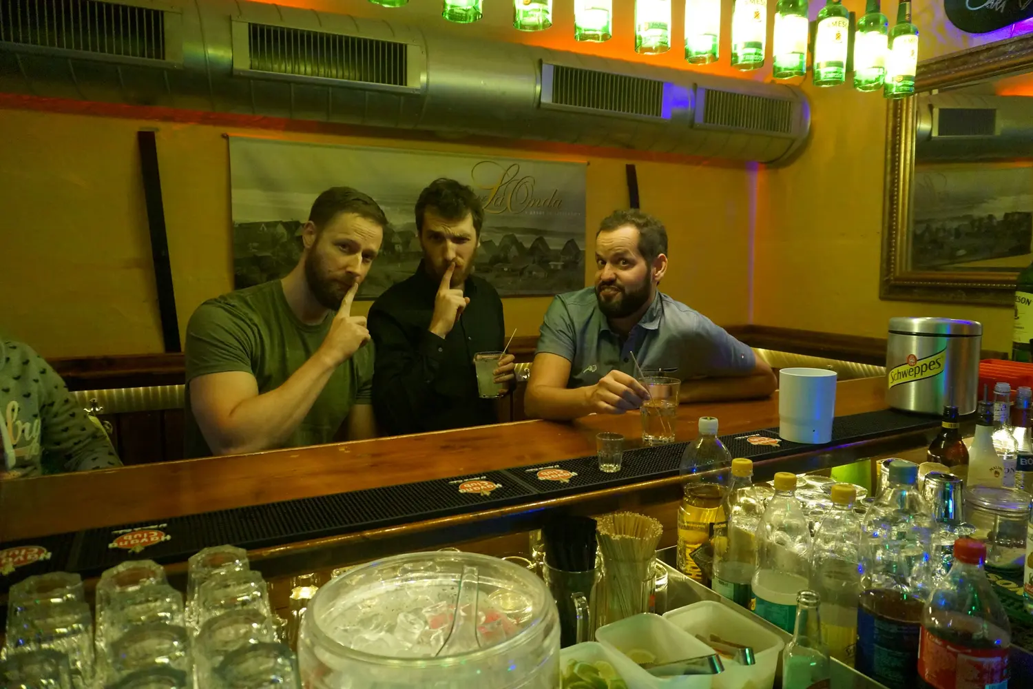 Three guests at the counter hold a finger to their lips, the bottle lamps above them