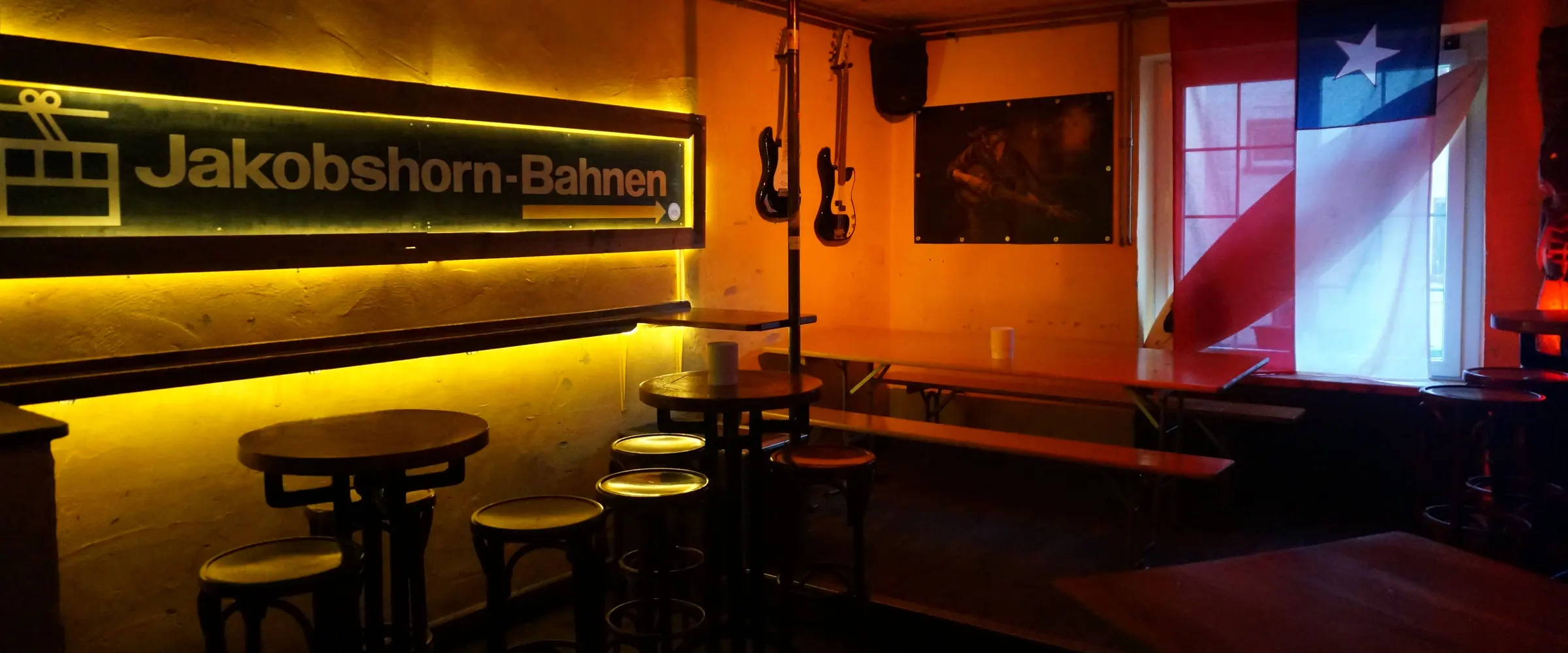 High tables and bar stools under the lit sign of the Jakobshorn cable car, two guitars on the wall, a flag at the window